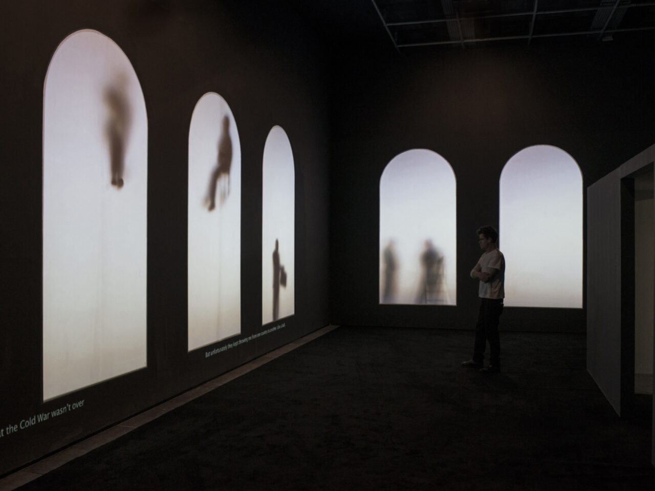 A dark lit room with five projected arches on the wall, with figures of people standing, leaning and floating, there are various passages of writing at the bottom of the wall.