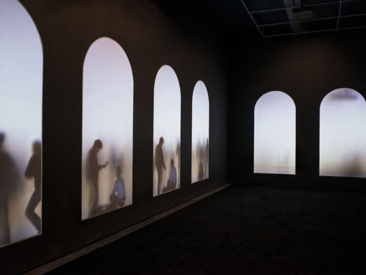 A dark lit room with five projected arches on the wall, with figures of people standing, leaning and floating, there are various passages of writing at the bottom of the wall.