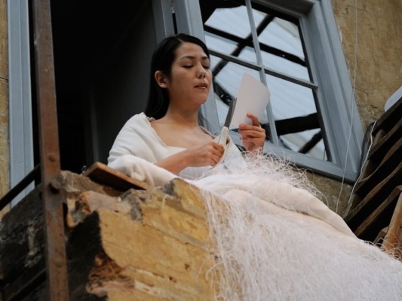 A person on a balcony, they are wearing all white cutting paper, the paper is trailing down with wire from the balcony to a sculpture in the middle of the warehouse.