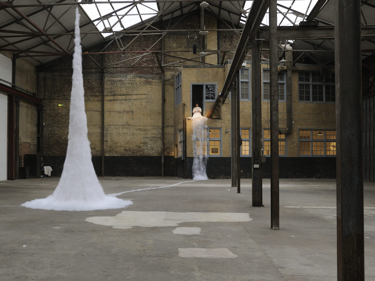 A large warehouse with metal pillars and a yellow brick wall at the back. In the space is a person on a balcony, they are wearing all white cutting paper, the paper is trailing down from the balcony to a sculpture in the middle of the warehouse. The sculpture is a fountain shape made of white paper and wire.