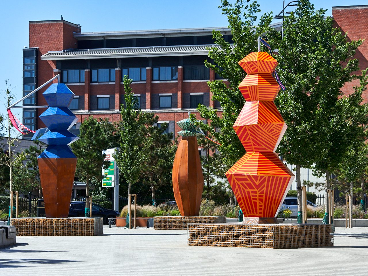 Three totems on Princes Dock, one geometrically painted red and yellow, one painted blue and the bottom half is covered in rust and the last is almost entirely covered in rust except for its green top