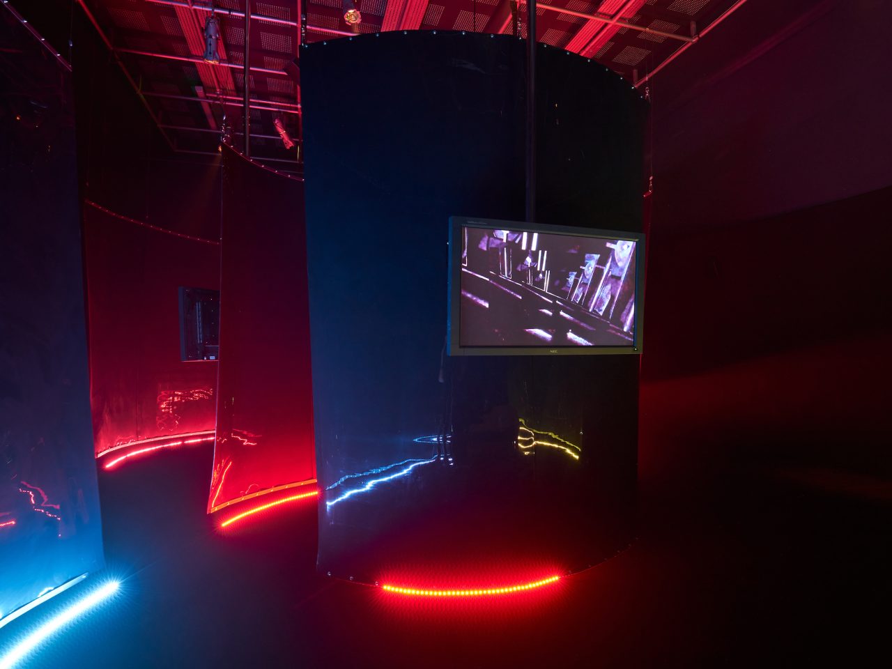A mixed media installation with multiple red and blue fabric screens with red and blue led lighting underneath. There are TV's in-between the sheets of fabric.