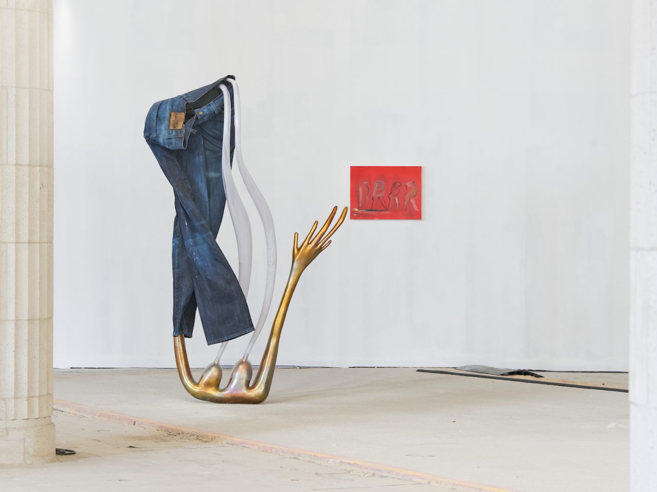 A tall sculpture with long gold arms reaching up with a hand pointing upwards. There are a pair of blue jeans draped over the top and coming out of the jeans are a milky white to clear plastic, shaped as if it were a pouring liquid.