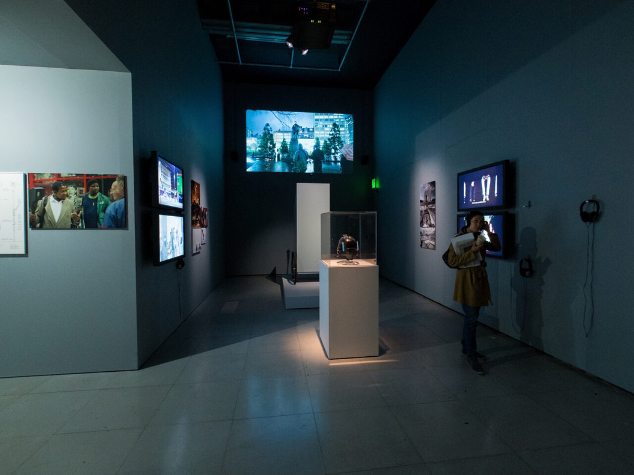 A room installation of artworks, there are four screens, one projection, mounted photographs and graphs and objects on platforms in the middle of the space.
