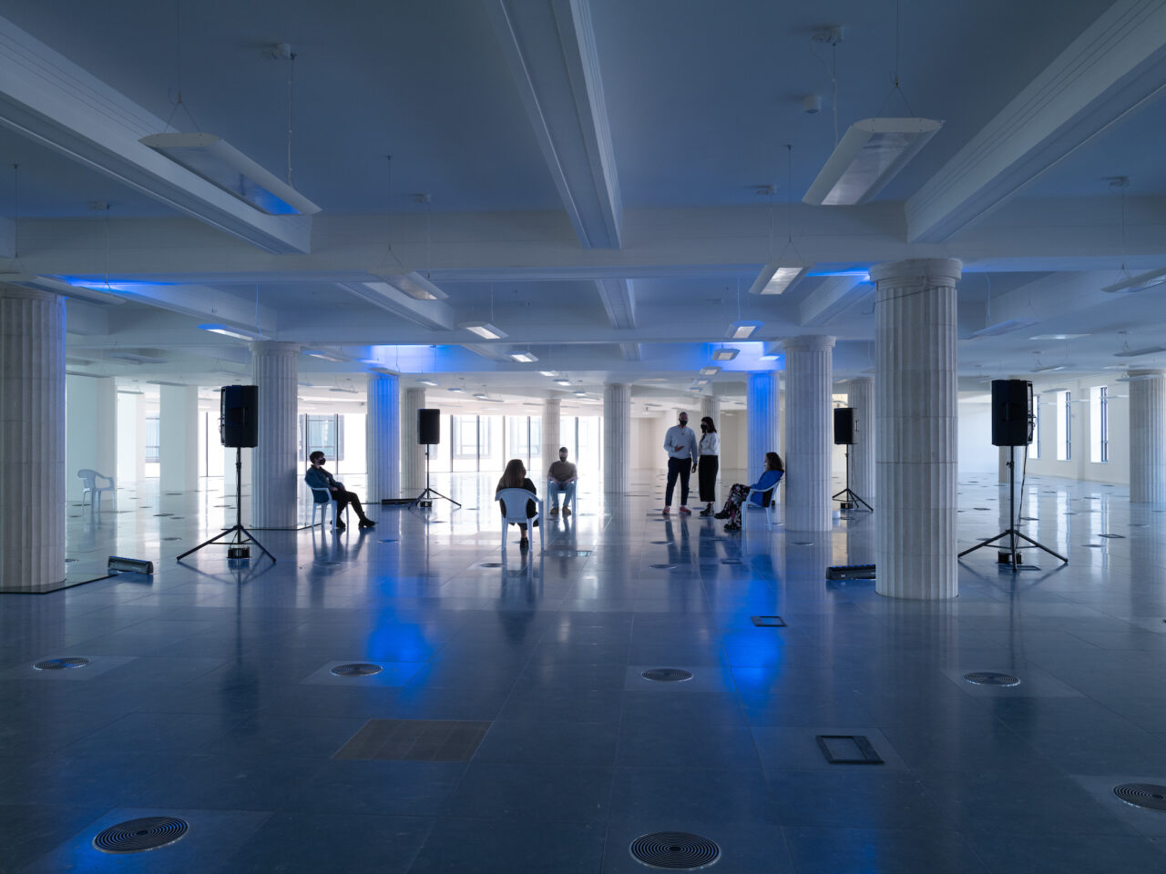People in a large white room in the Lewis's Building with large white pillars throughout. In the middle of the room are speakers on stands all facing each other, the middle space is lit up with blue lighting and there is a sound piece playing.