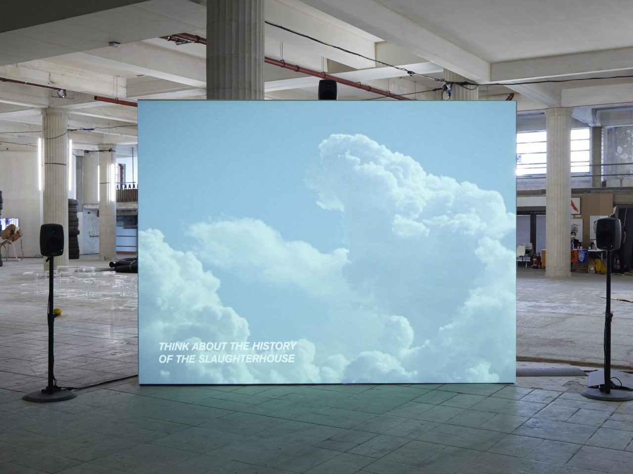 A free standing projector screen with a film still of clouds and white text reading, 'Think about the history of the slaughterhouse'