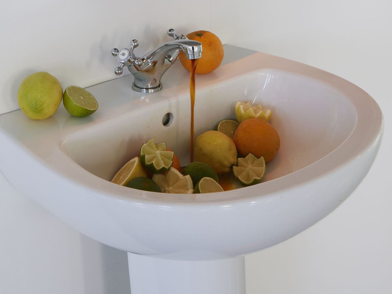 A sink with lemons, limes and oranges, some full and some cut with a pattern. They are in the sink with the tap letting out an orange liquid.
