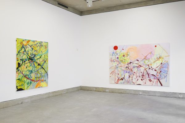 Two large abstract paintings on a white wall, the painting on the left is mostly yellow with blue markings and the painting on the right is mainly light pink with yellow, red and blue markings.
