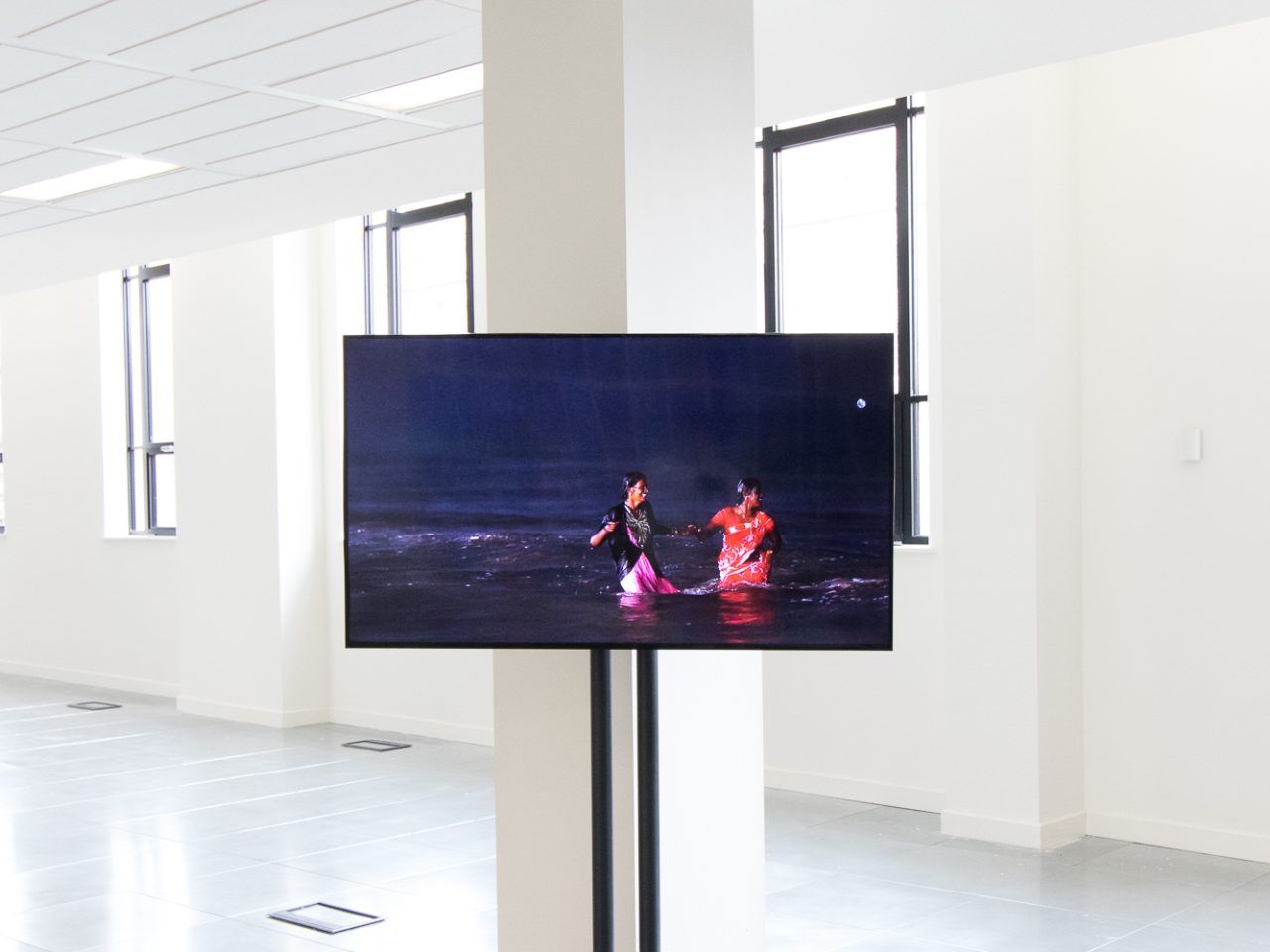 A Television Screen on a stand with a film playing of two people smiling in the sea while its raining.