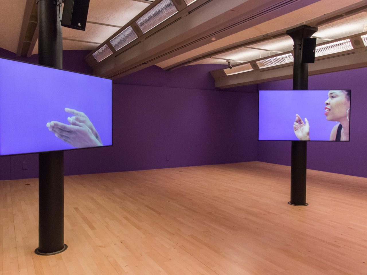 a gallery with purple walls and two screens. The screens show close up videos of the screens interacting with each other. The background of the videos, are the same purple as the gallery's.