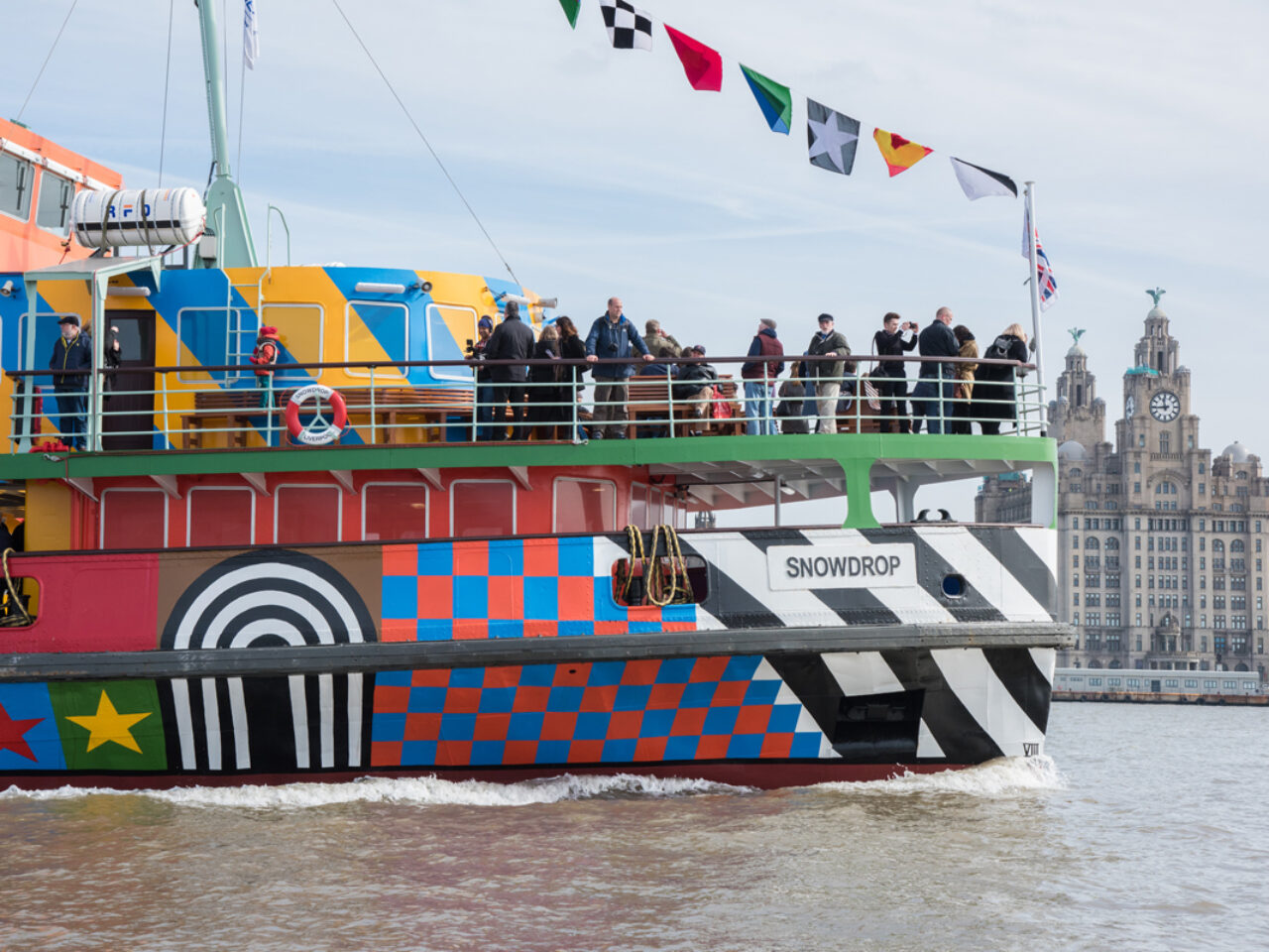 A colouful patterned ferry sailing across the River Mersey. There are people on the balcony looking out and you can see the Royal Liver Building in the distance