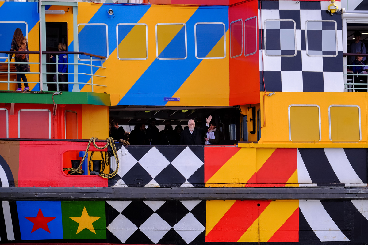 A a close up shot of a colouful patterned ferry. The artist is on the ferry and waving to the camera