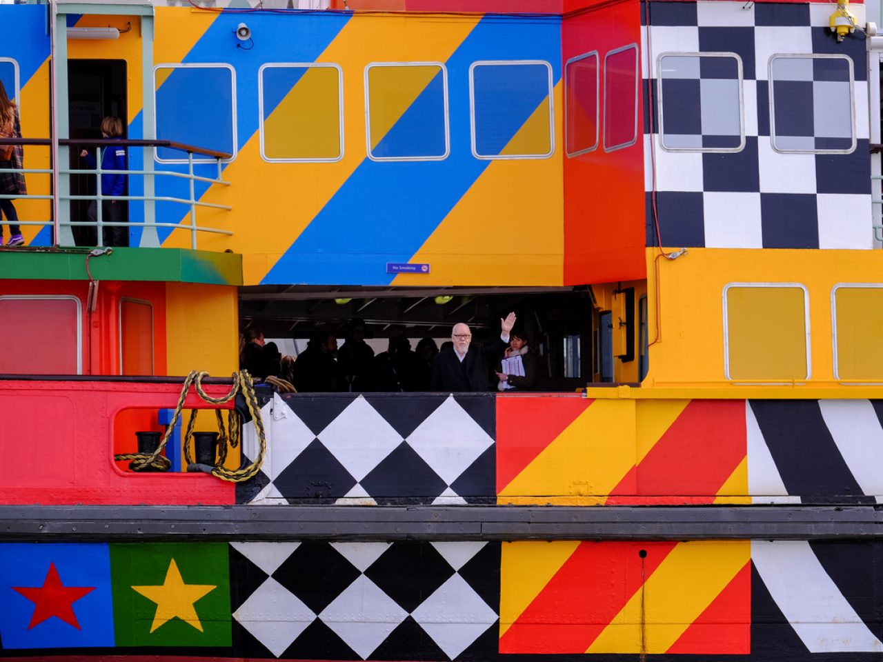 A close up shot of a colouful patterned ferry. The artist is on the ferry and waving to the camera