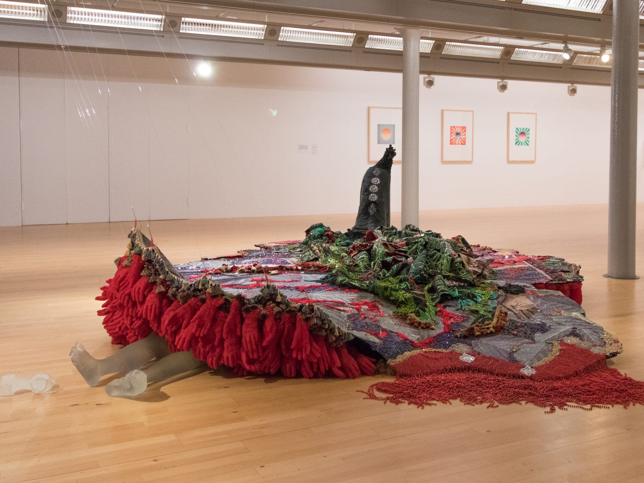 A platform made up of purple, green and red fabric and beads. Underneath are stacked red gloves, lined with tassles. Coming from underneath are a persons legs lying down with their shoes coming off, the legs and shoes are made out of frosted glass.