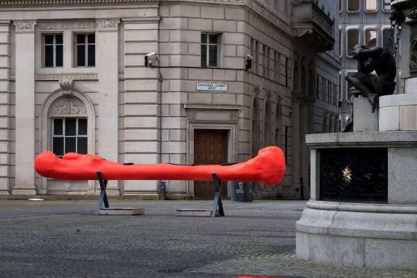 A installation within Exchange Flags, there is a bone shaped canoe, it is bright red and sat on a metal stand.