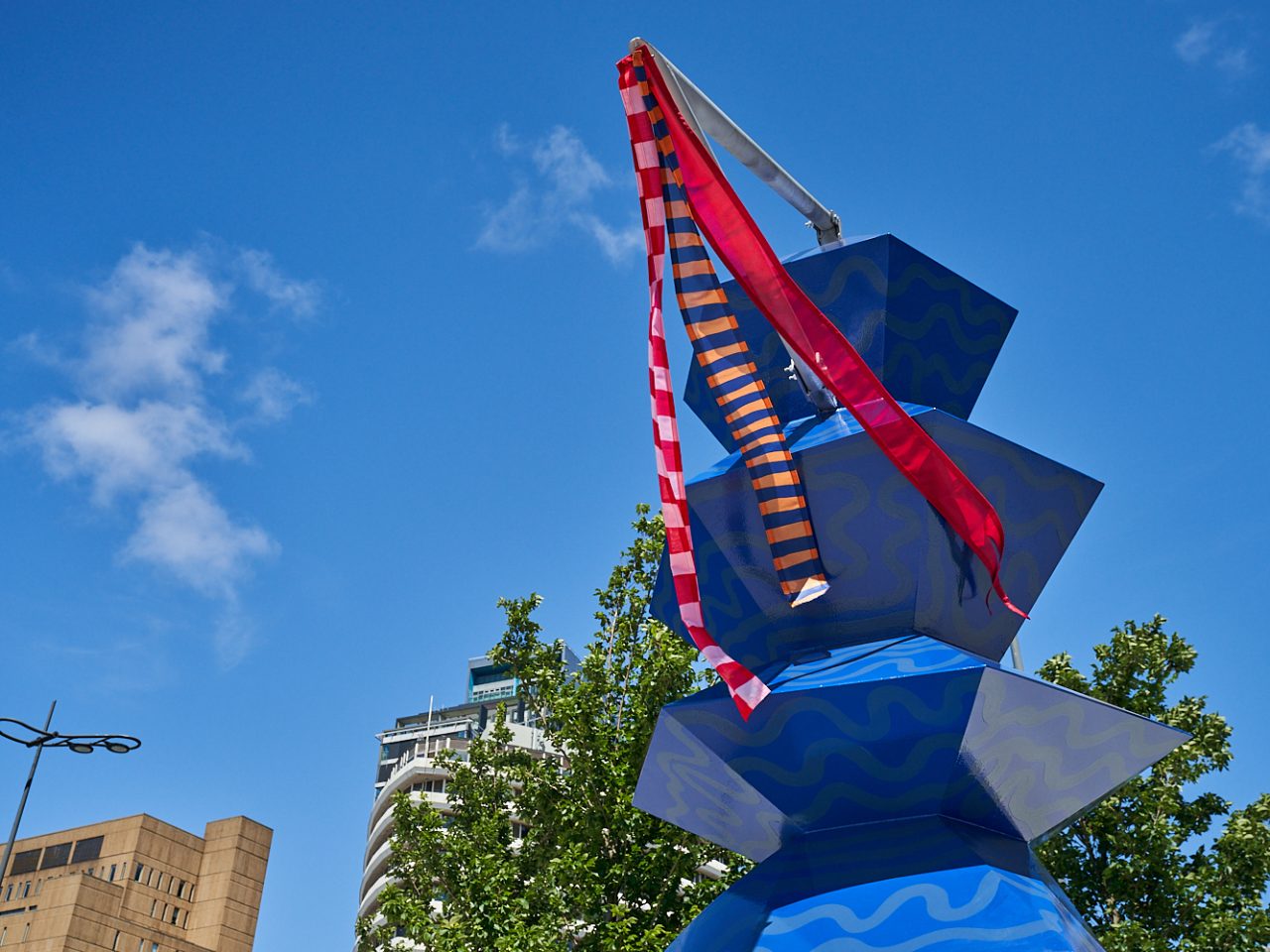 A totem painted blue with pale blue squiggles. At the top of the totem is a pole pointing out of it and 3 ribbons moving in the wind