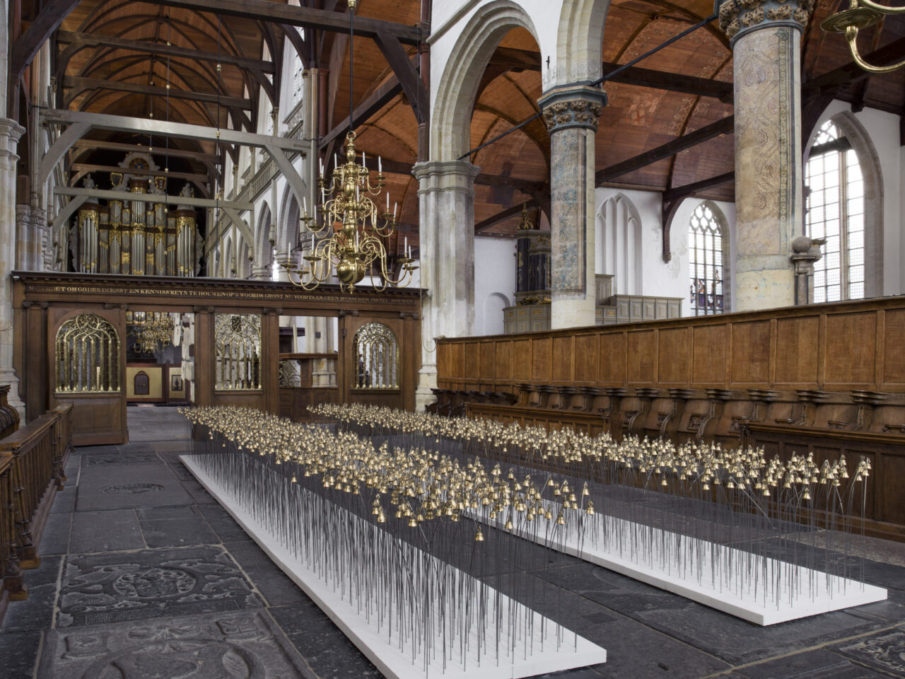 An installation of 2 long pathways of bells. It has a walkway in the middle so you can walk through it, this installation is inside a church⁠