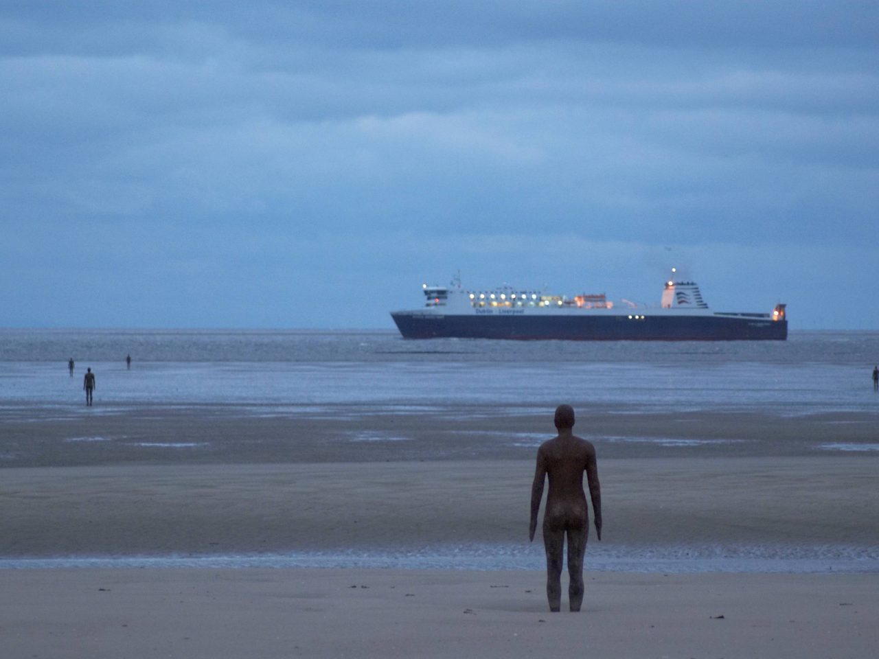 A beach scape with multiple life-size cast-iron moulds of the artists own body. There is a ship sailing within the water.