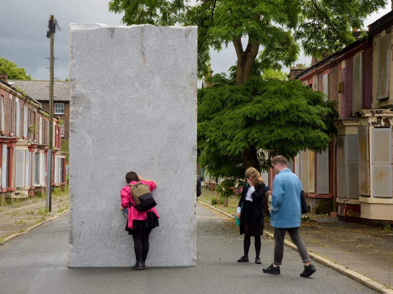 3 people standing around a sculpture. The sculpture is a huge granite boulder and is situated in the middle of a road of terraced houses