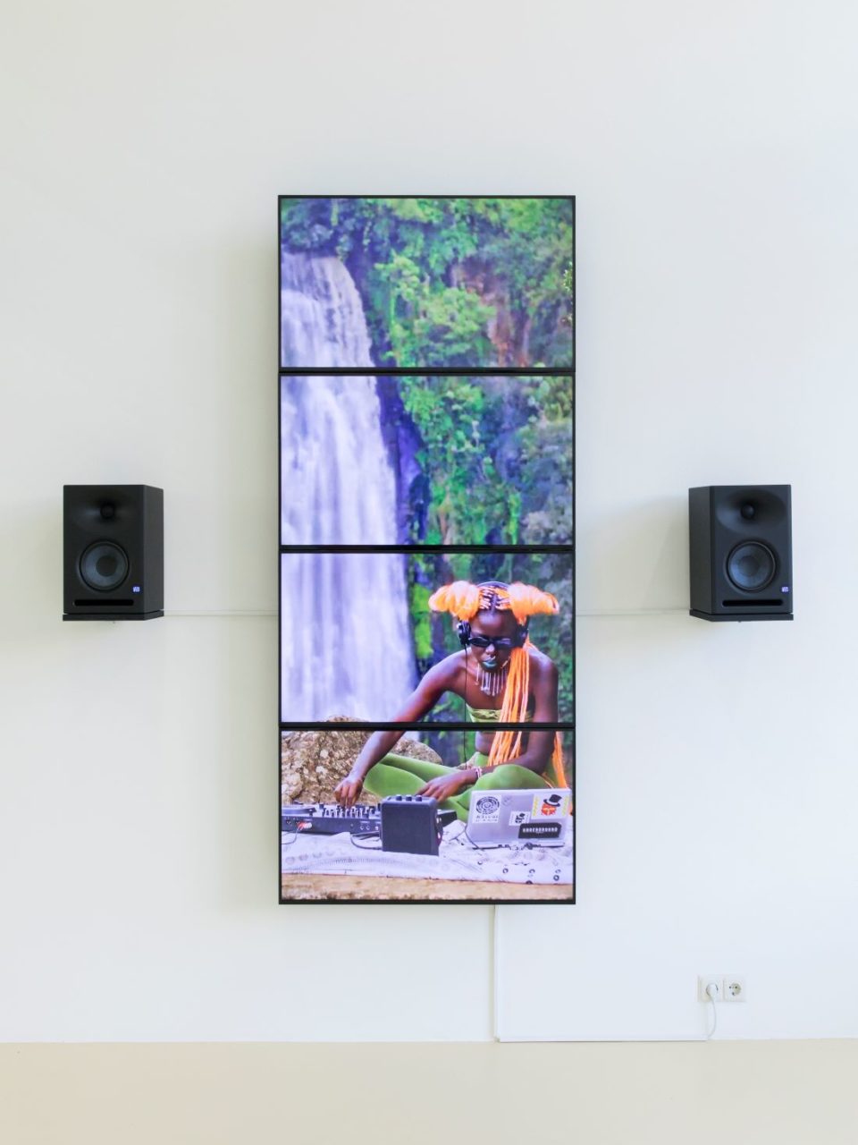 Four medium screens connected vertically with speakers either side of them. The screen shows a person DJ'ing, they have neon orange hair. In the background is a waterfall.