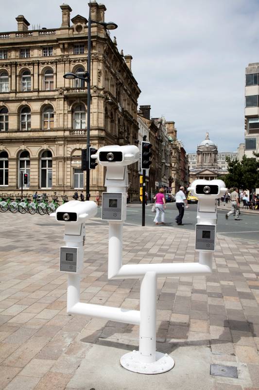 An installation which painted white tubular metal pipes with three telescopes at varying heights. The installation is within a city centre with people walking around
