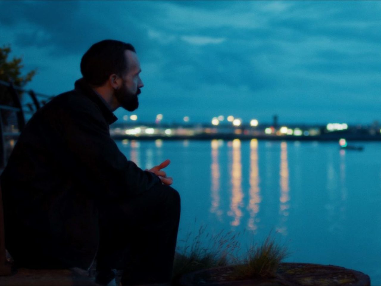 A film still. The actor is sat on the side, looking out to a lake, it is starting to go dark.