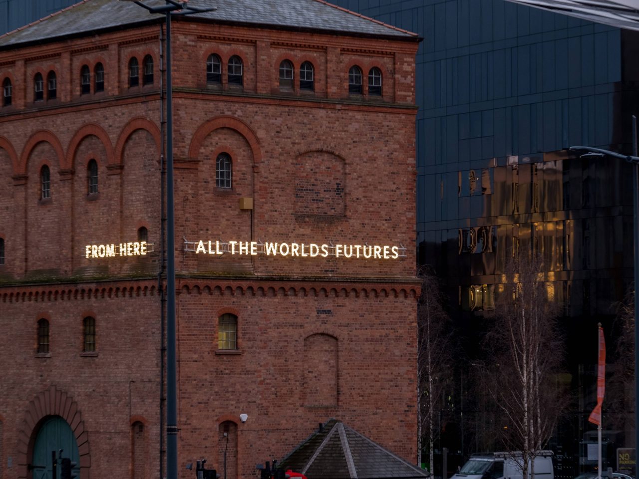 A close up of a building, the building has glowing lights reading 'from here, all the worlds futures'