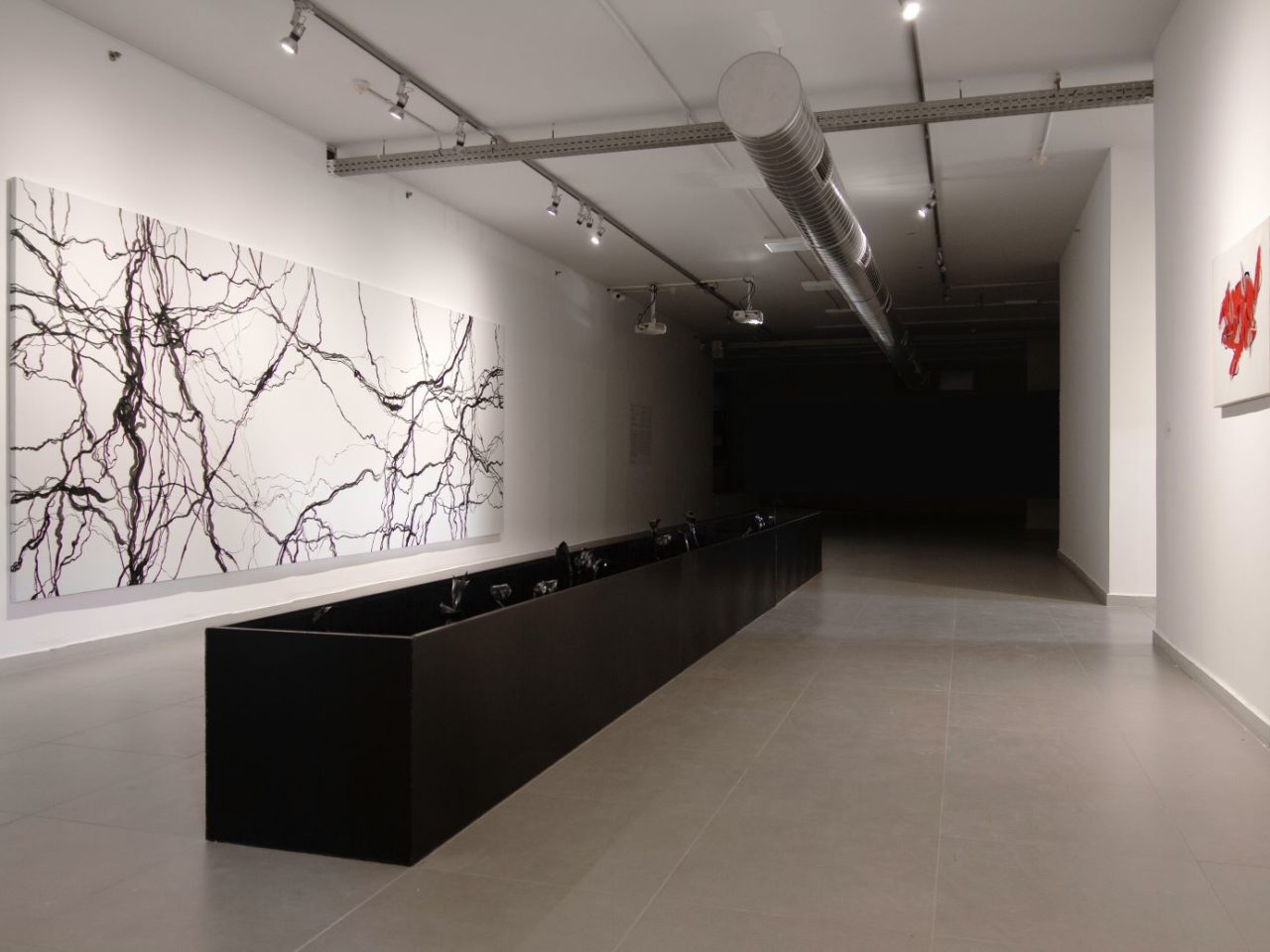 In a white gallery space, there is a large, long painting on the wall. The painting has a white background and black lines that bend and curl irregularly. In the middle of the Gallery space is a black case, longer the length of the painting.