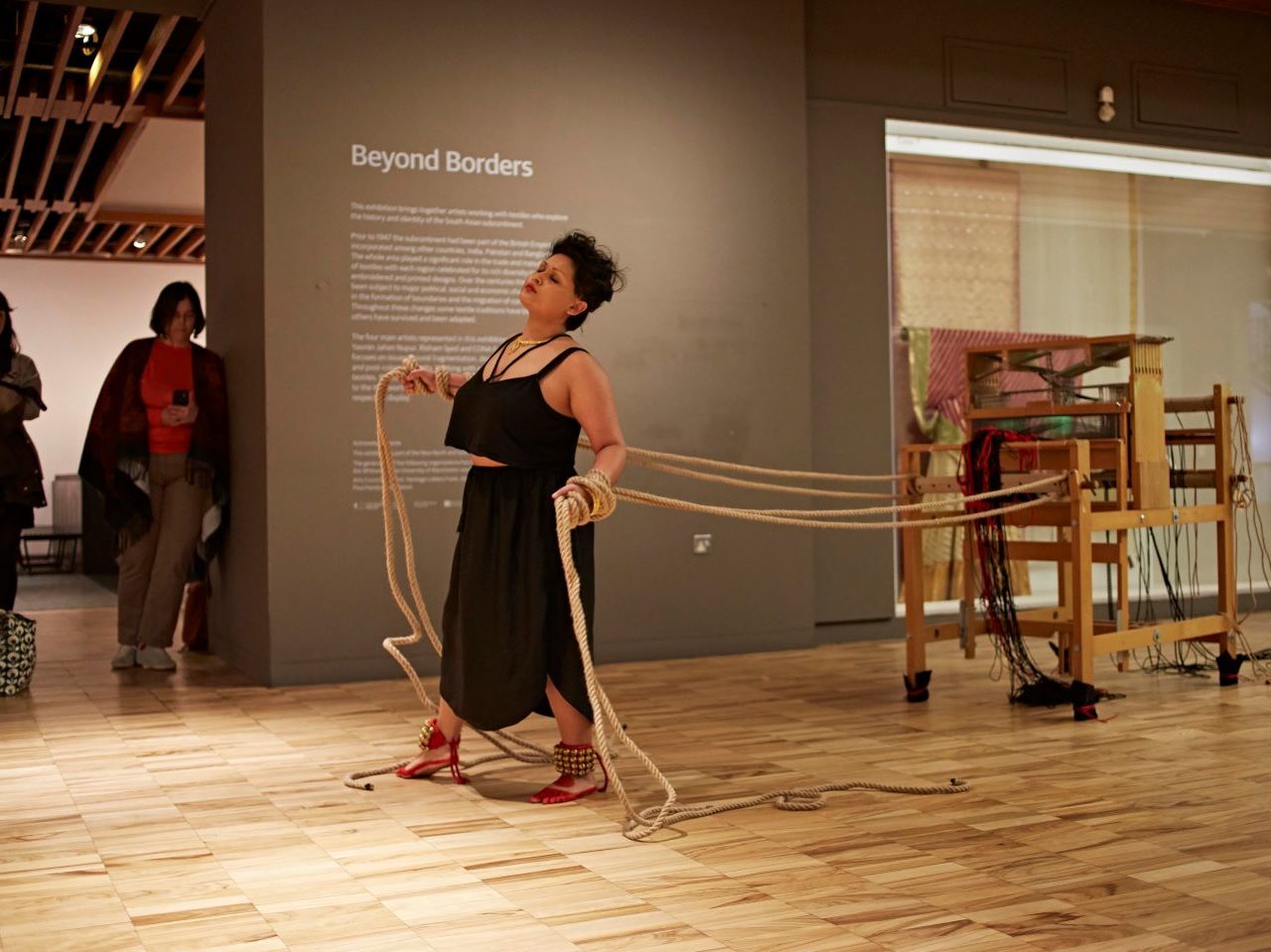 The artist performing in a gallery space, they are attached to a loom through ropes. They are wearing a long black skirt and a black top, their feet are painted red and they have bells around their ankles.