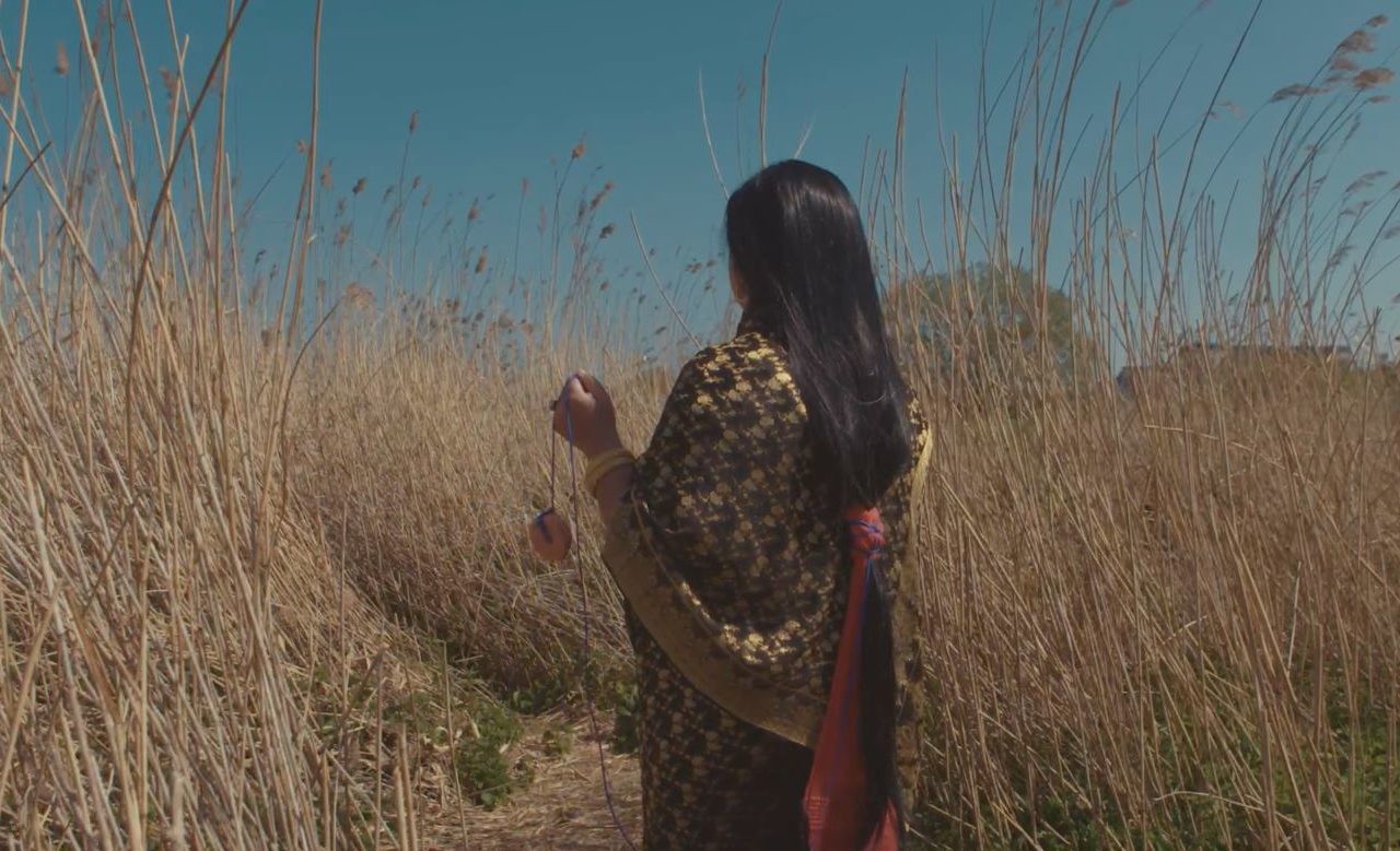 Someone standing in the middle of the dunes, the surrounding Marram grass is grown taller than the person standing. The person has their back to the shot of the camera, they have hip-length black hair and are wearing a patterned shall and skirt.