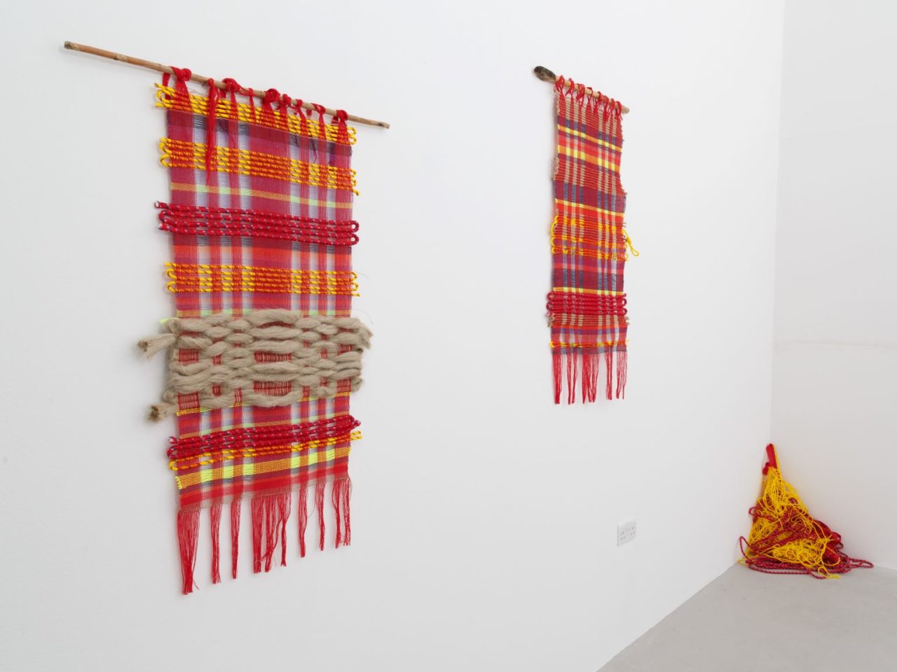 Two textile tapestries, hanging on a wall, they are red with hints of yellow and blue. In the corner of the room is a pile of lose thread coloured red and yellow.