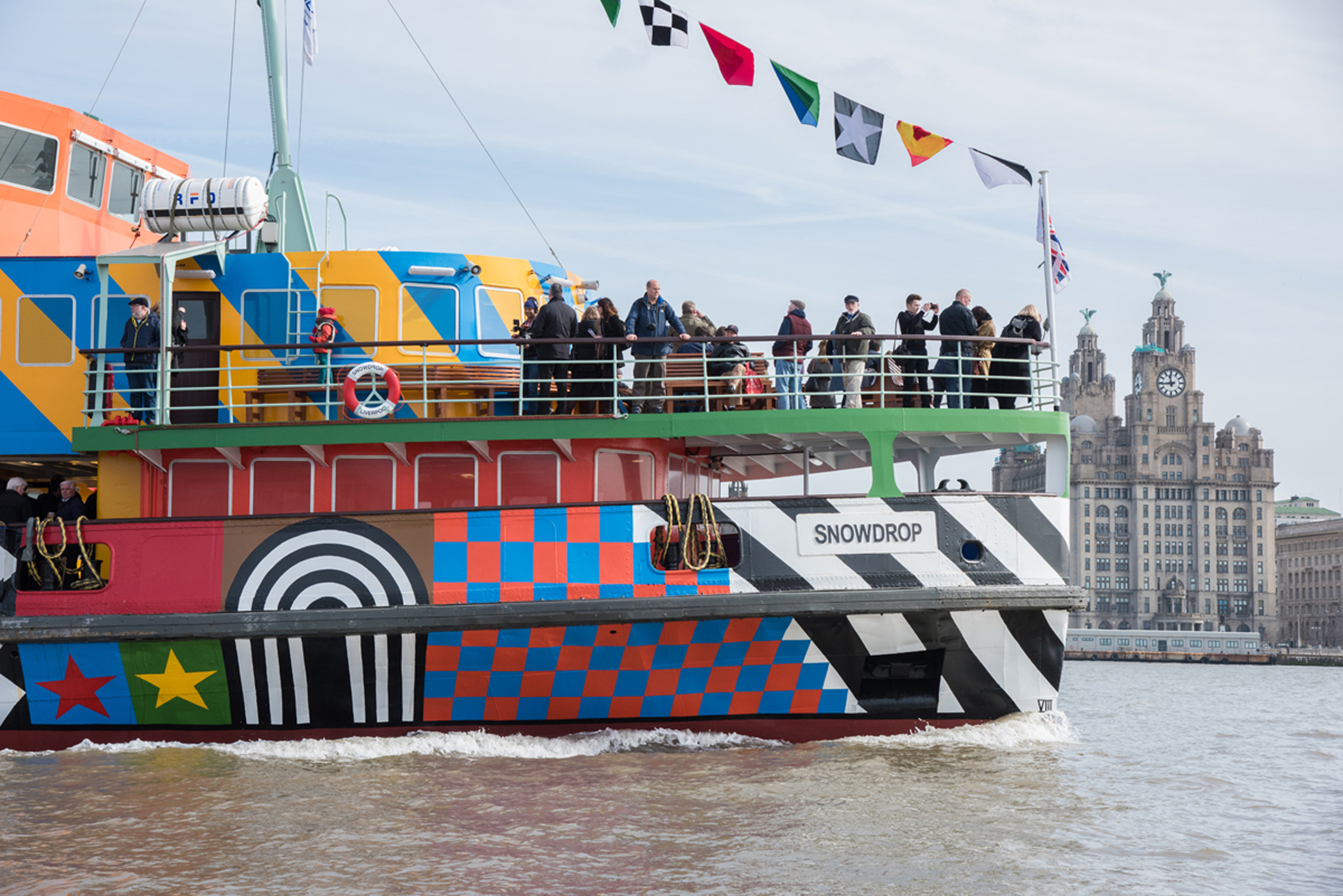 A colouful patterned ferry sailing across the River Mersey. There are people on the balcony looking out and you can see the Royal Liver Building in the distance
