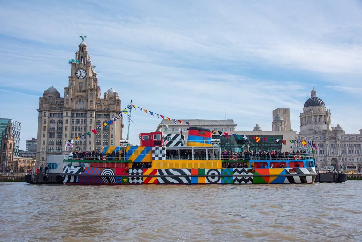 A colouful patterned ferry sailing across the River Mersey. In the background is the cityscape of Liverpool
