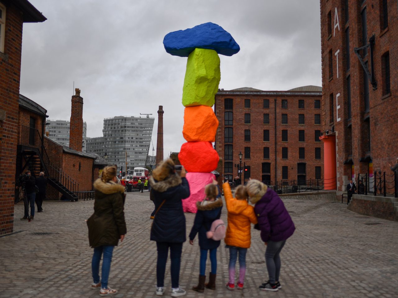 A family of 5 pointing and take a picture of 10-metre high sculpture on Liverpool’s waterfront, the sculpture consists of 5 vertically-stacked rocks painted in bright fluorescent colours