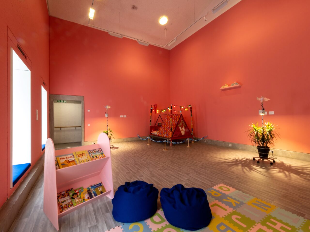 An installation including plants, hand-sewn pastel-coloured bats, both hanging and on a shelf. There is a space with blue beanbags and a foam floor. In the corner is a colourful tent, that is mostly red and has colourful lights hanging above it