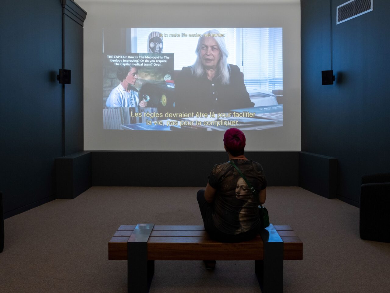 Someone sat on a bench in a dim lit room, they are watching a film that is projected onto a white wall.