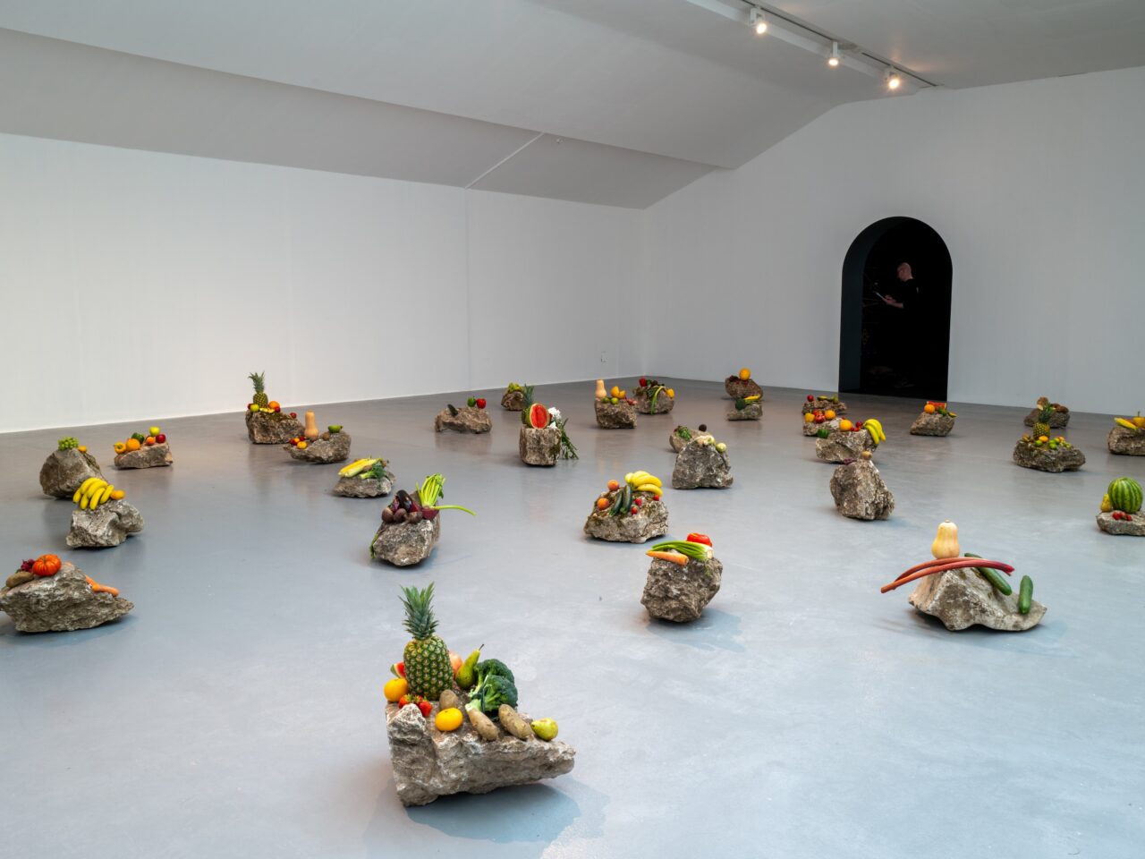An installation in a white gallery space. The installation is spread along the whole gallery floor and consists of large rocks with a variety of fruit laid upon them