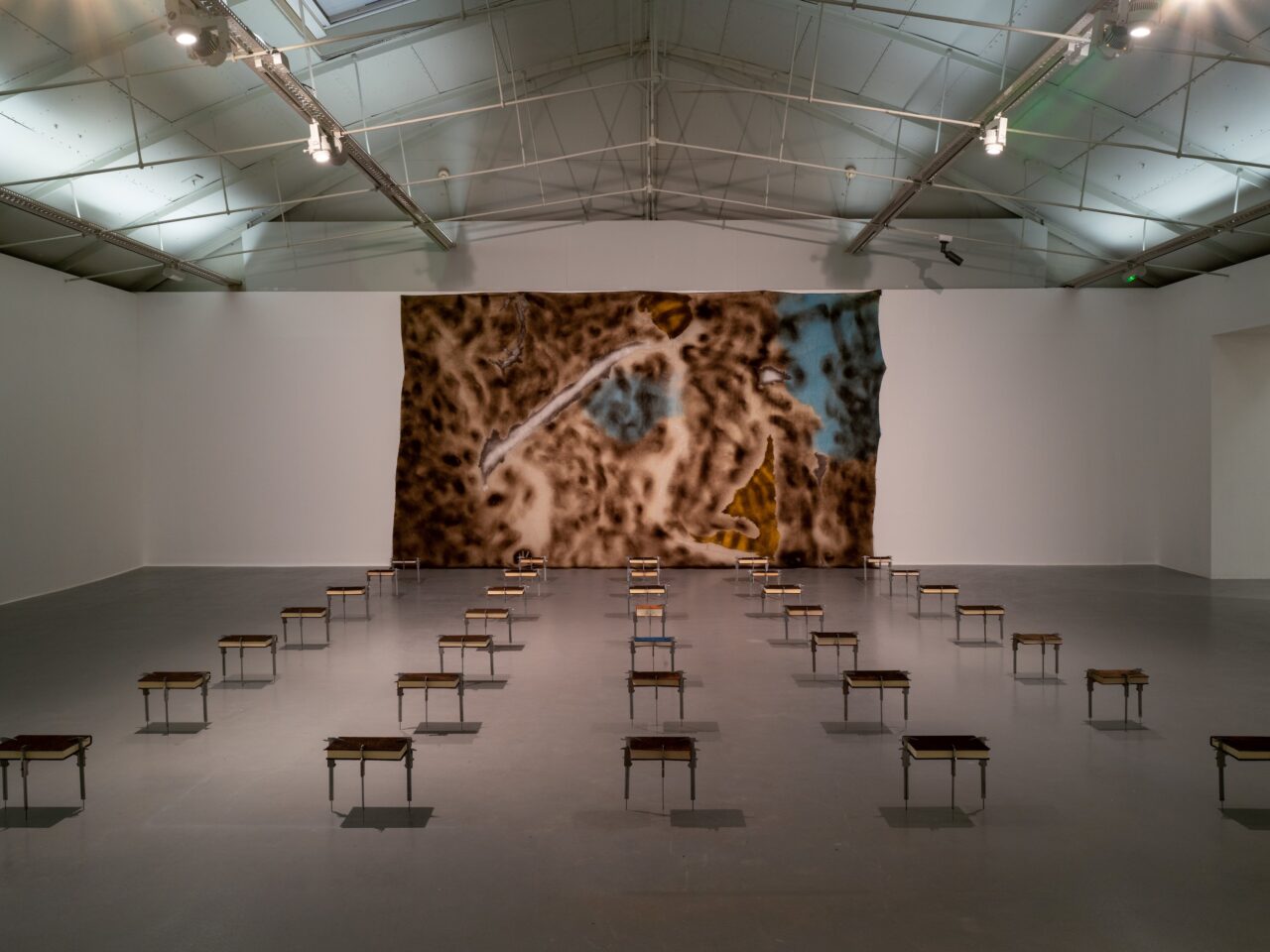 An installation, on the back wall there is a large wall hanging that is marked with coffee grounds and ink, its is mostly brown with aspects of blue. On the surrounding floor are books all held up by callipers. All but 2 of the books are covered in coffee grounds.