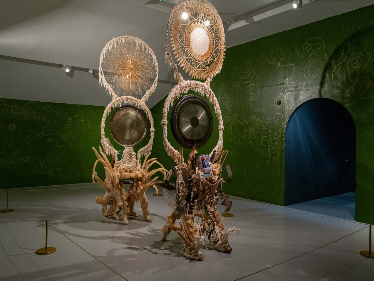 Two assemblies consisting of a gong, a mixture of steel, loofah and other mixed media materials, alongside objects collected from the artist on their travels. The surrounding walls are green with gold tracings.