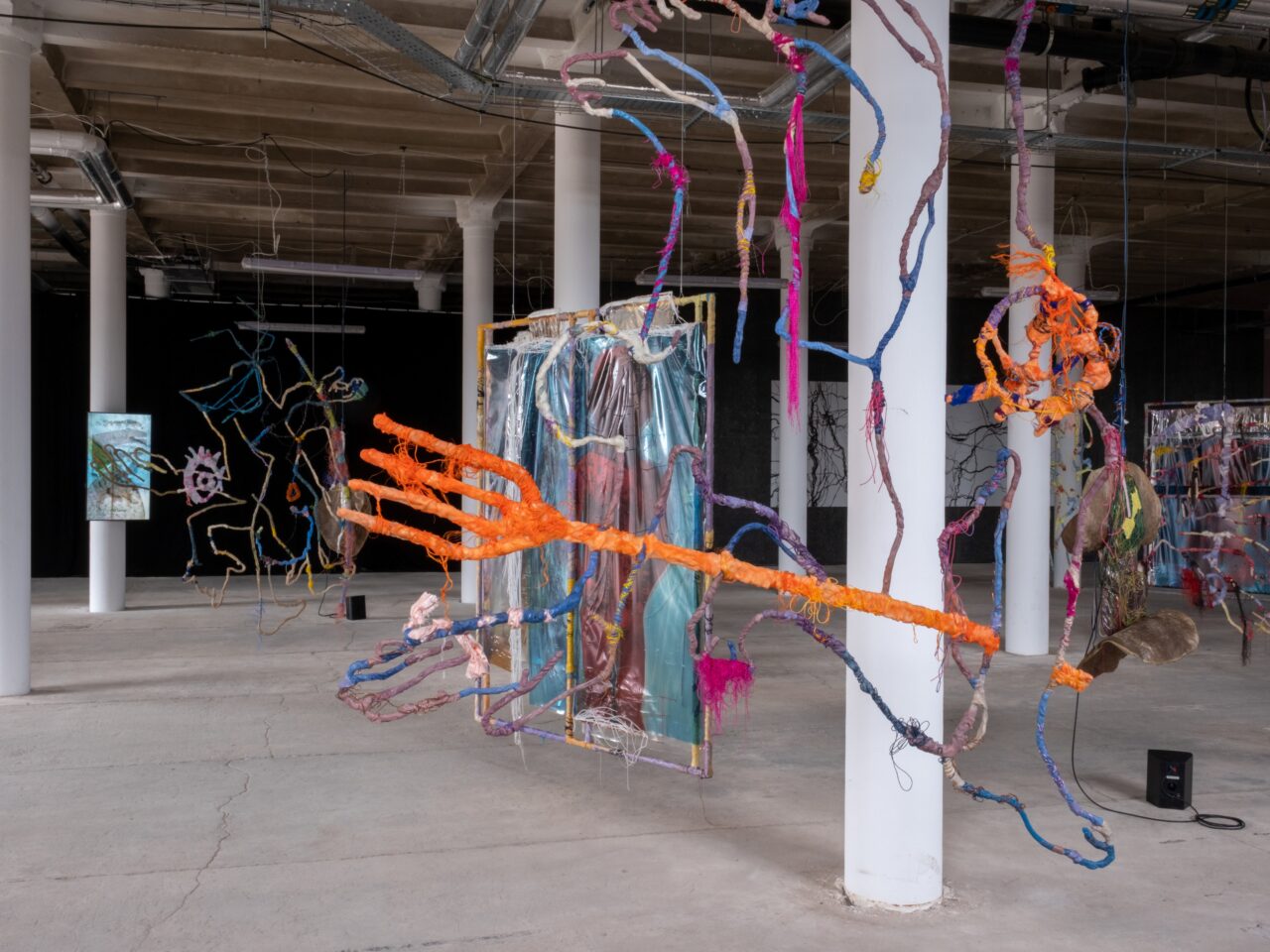 a large installation of hanging, textured mixed media sculptures and video work inside a brick warehouse. The various materials include fabric, UV print on vinyl and thread.