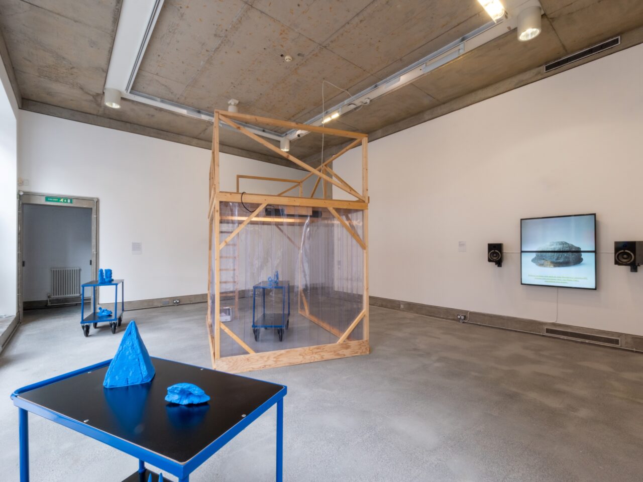 a gallery installation with white walls and grey floor. There is a black table with small blue sculptures. In the background, there is a video playing on a TV. In the middle of the room is a wooden framed structure with an enclosed plastic area.