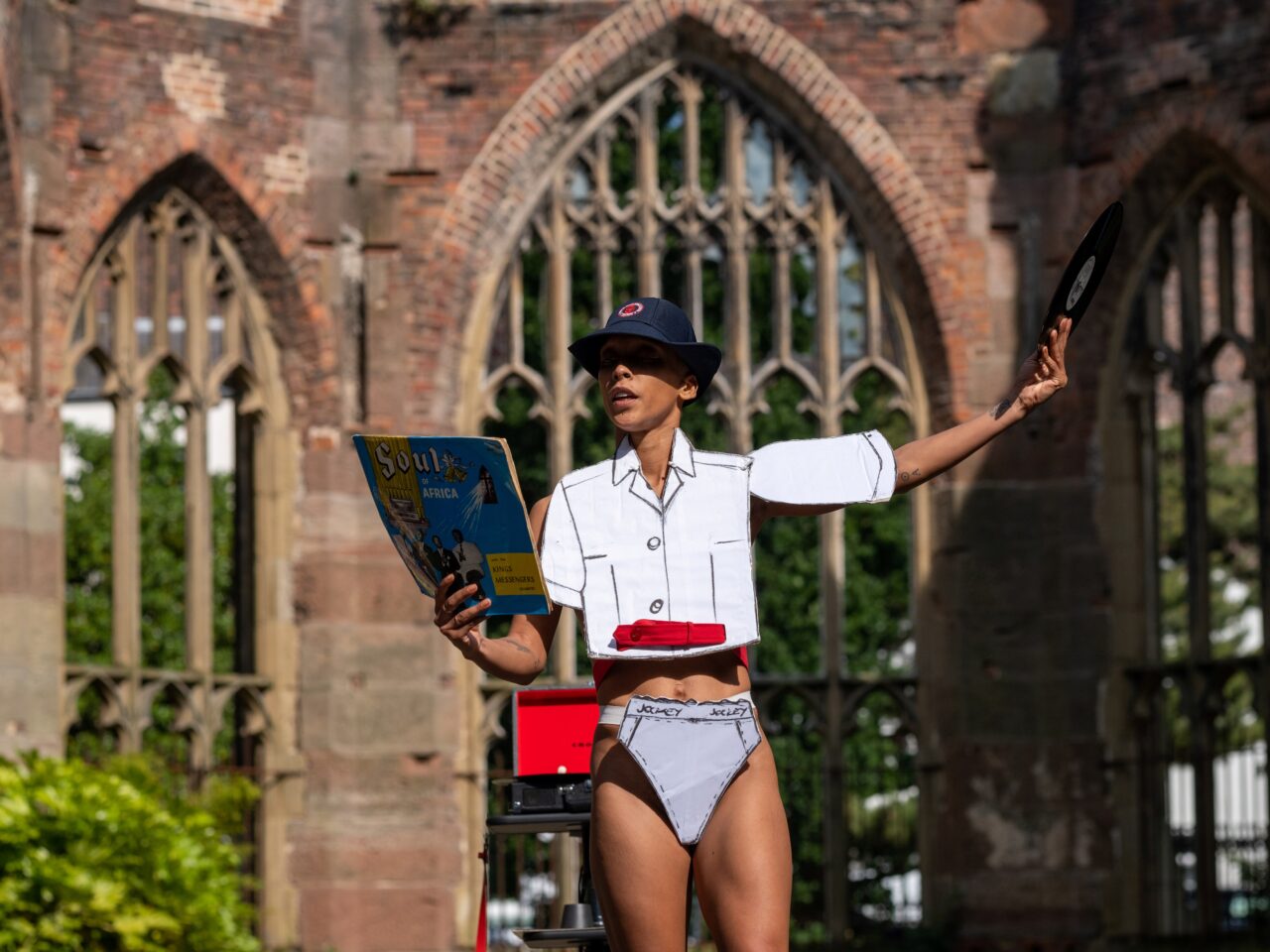 The artist presenting a live work in Liverpool's Bombed Out Church. They are wearing cardboard cutouts of a white school uniform and a hat. They are on a stage talking and with one hand holding a vinyl and the other holding the blue cover.