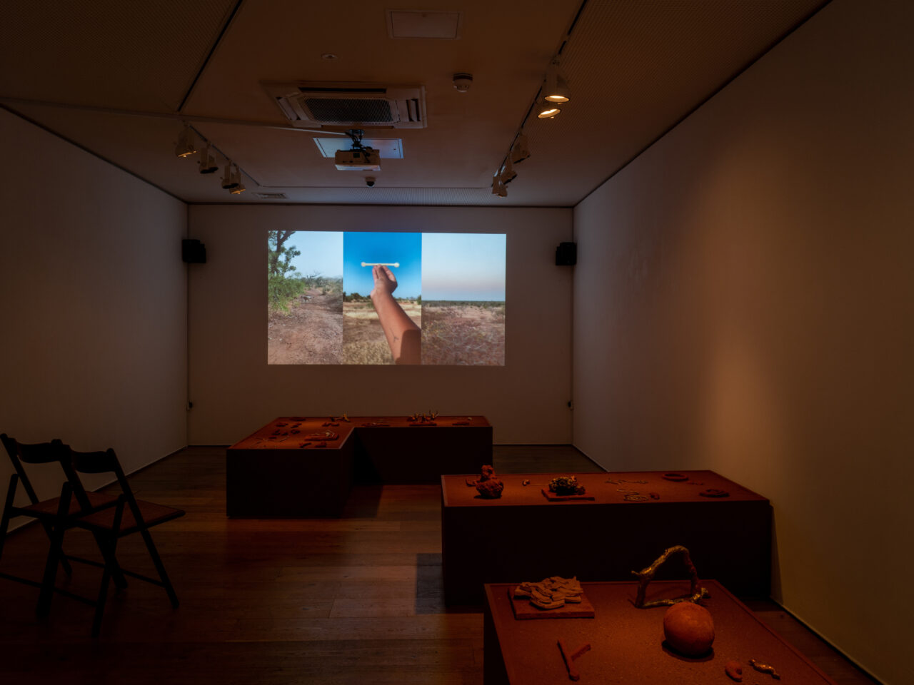 A mid-lit gallery space with a projection on the back wall with a split screen film. In the middle of the space is 3 large built plinths, which consists of mixed media and objects all in a orange sand-like colour and texture.