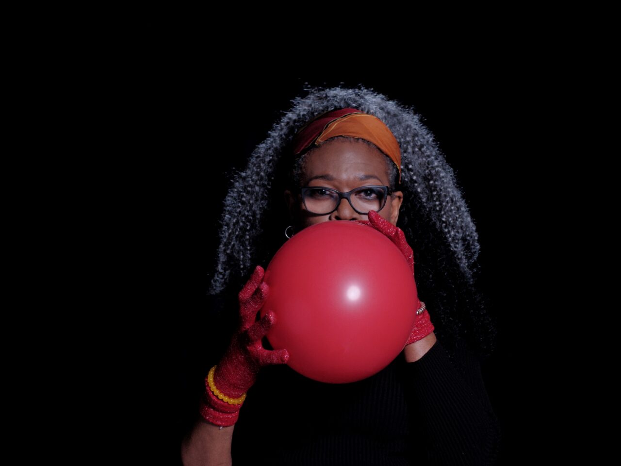 A video still of someone blowing up a red balloon, the balloon is in front of their face, the space behind them is black.