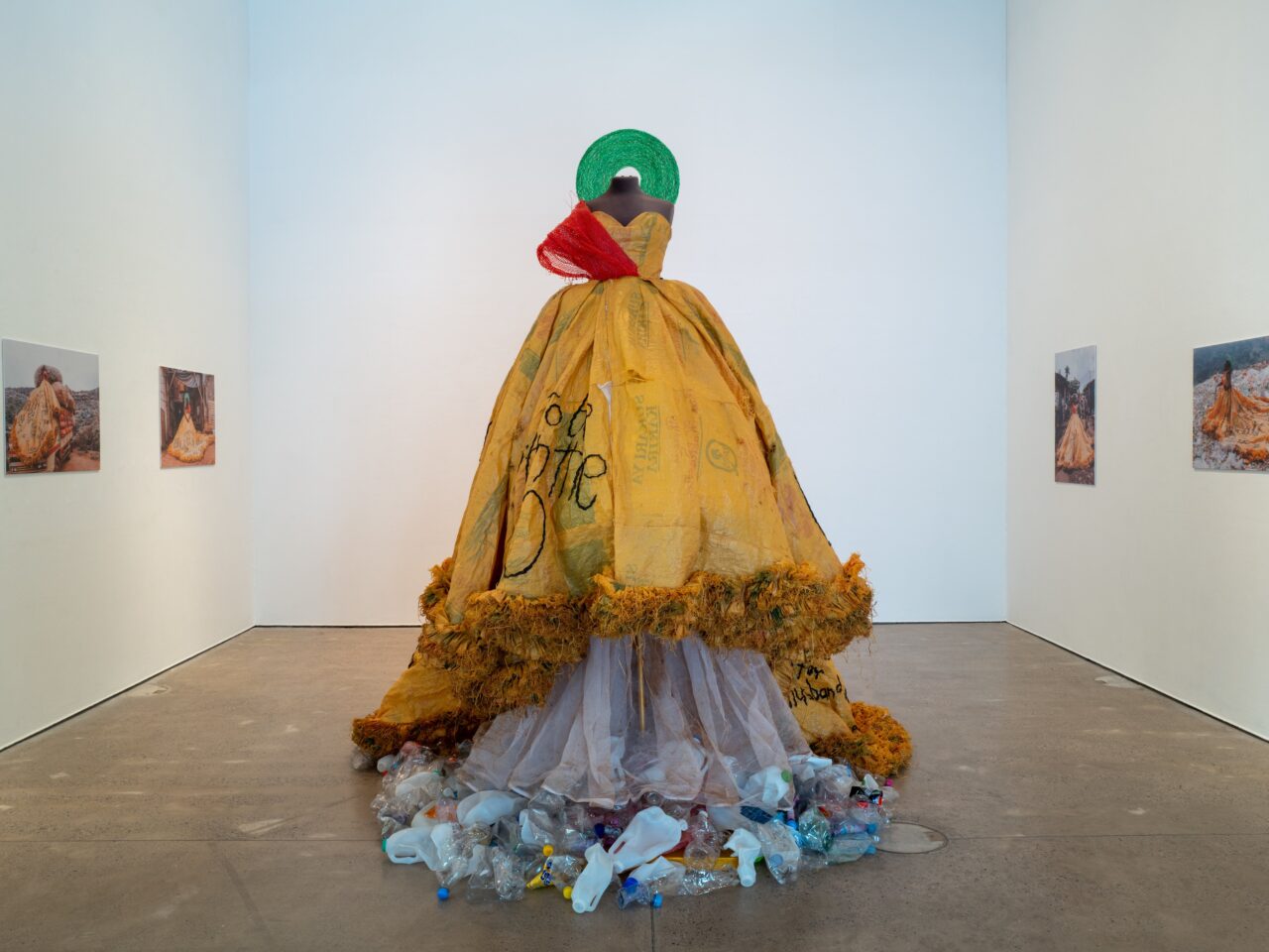 A white gallery space with a long yellow gown made with recycled materials in the centre of the floor. On the white walls are photographs of the artist wearing the gown in their hometown.