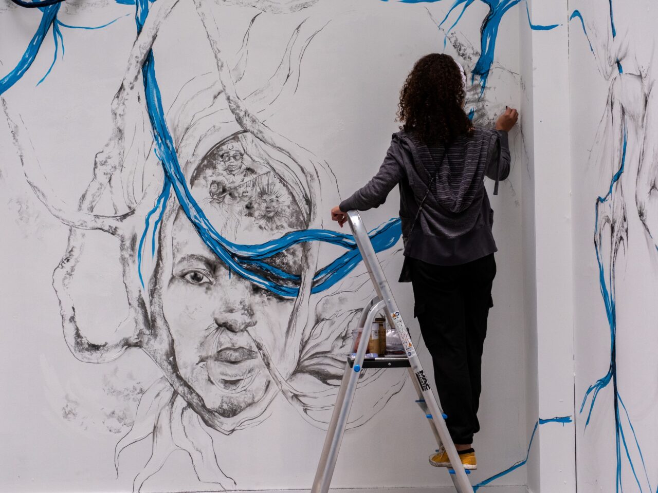 The artist Shannon Alonzo, standing on a ladder, wearing headphones and drawing with graphite onto a wall, creating a site-specific mural.