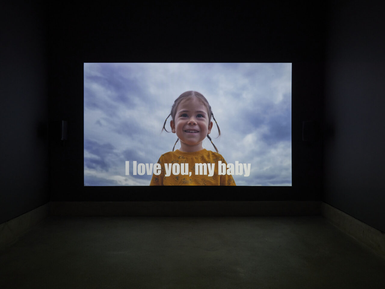 a dark room with a film projection, the film is of a young child looking at the camera. At the bottom of the film it reads ‘I love you, my baby’