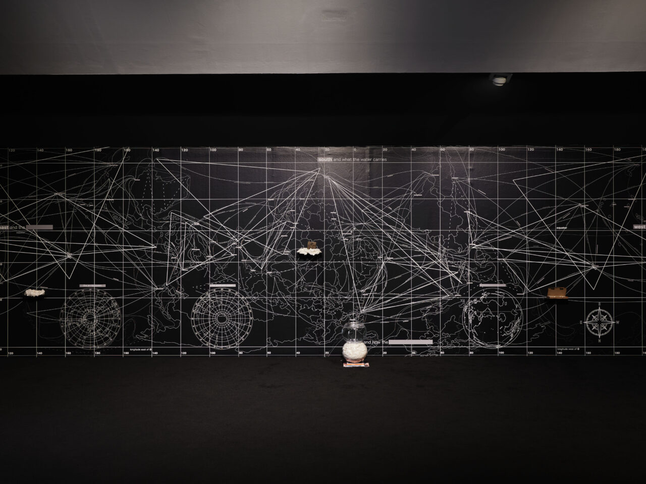 An installation with a large vinyl sticker covering the entire wall. The vinyl is black with white mapping and graphs covering it. There are shelves with various objects on them within the wall.