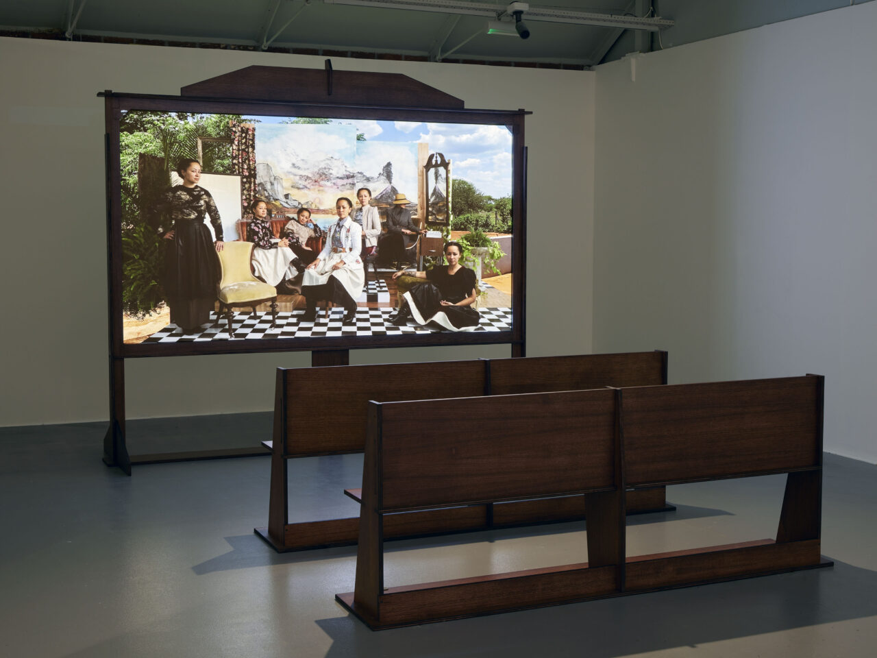 a projection screen which consists of the artist as seven different characters, in front of the screen are bespoke benches, made to mimic a Victorian design