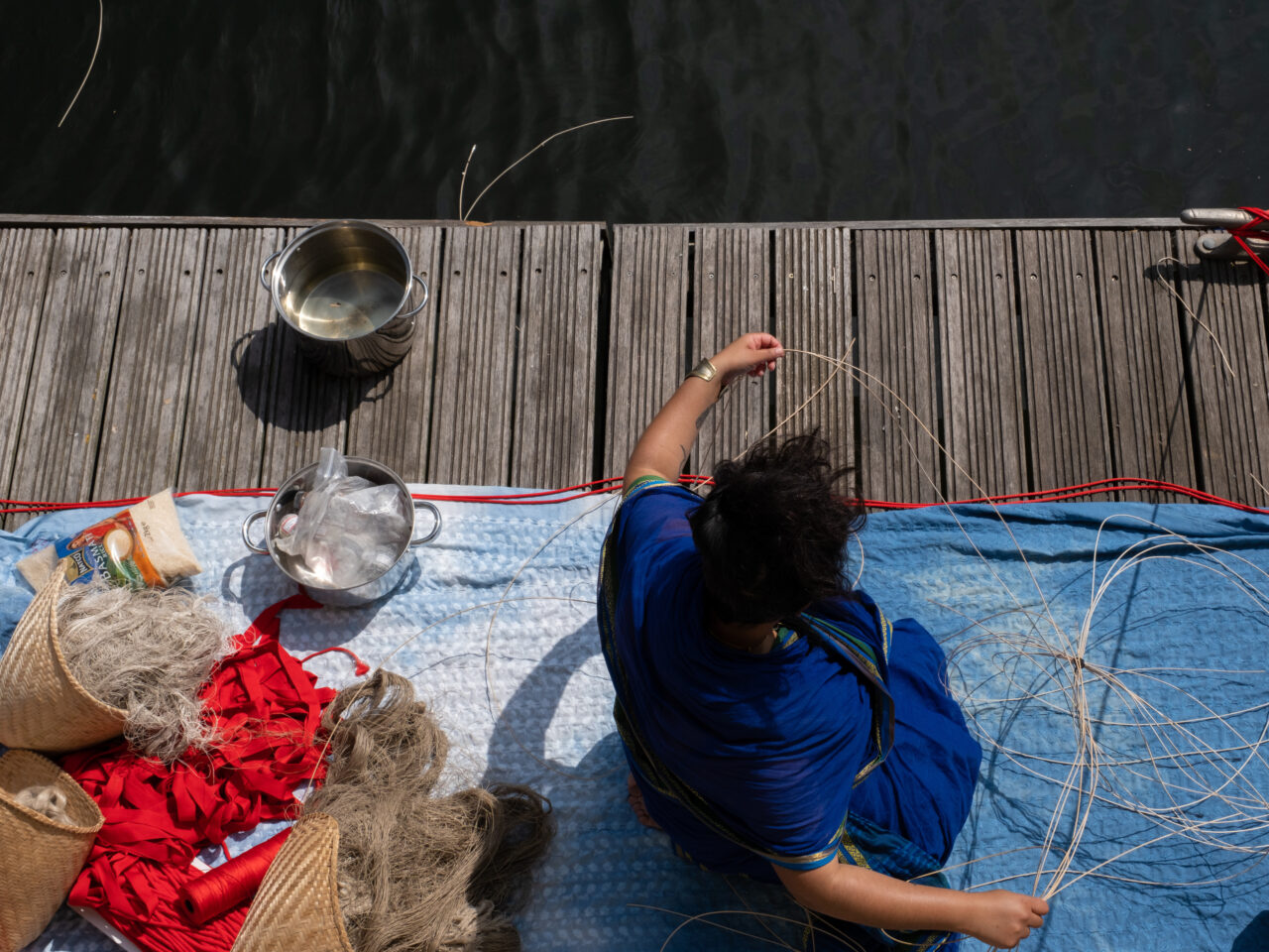 The artist is sitting on the wooden dock, they are wearing blue. Around them are woven baskets, textiles and a metal vessel.